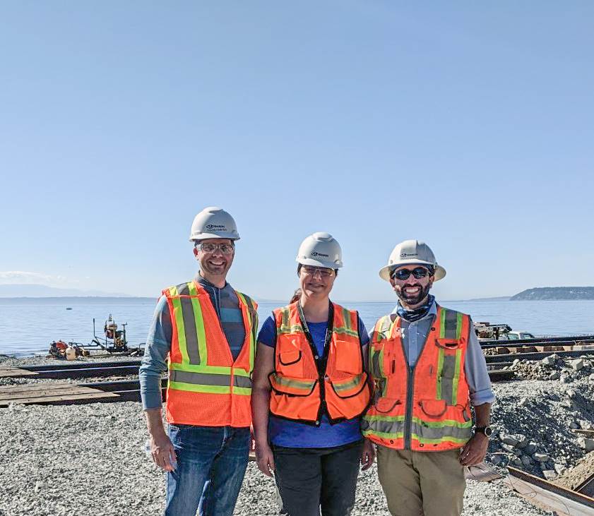 Three people in hard hats, safety vests and safety glasses stand in front of a rail bridge under construction with Puget Sound in the background