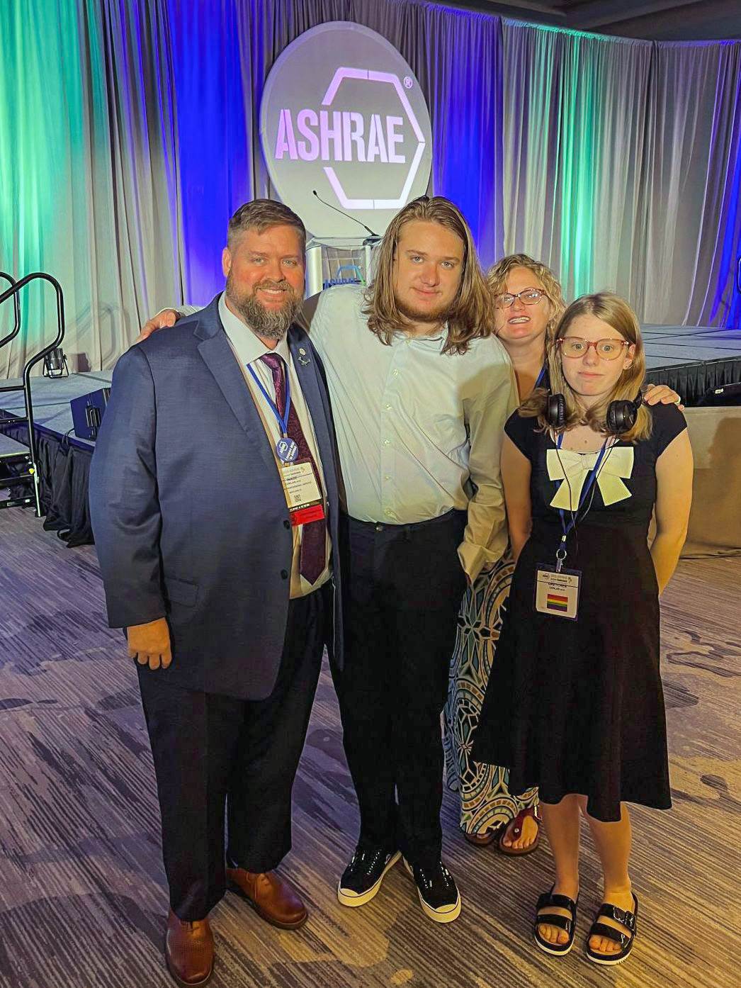 A family of four stands in front of a stage with a lectern and an ASHRAE logo behind it a hotel ballroom