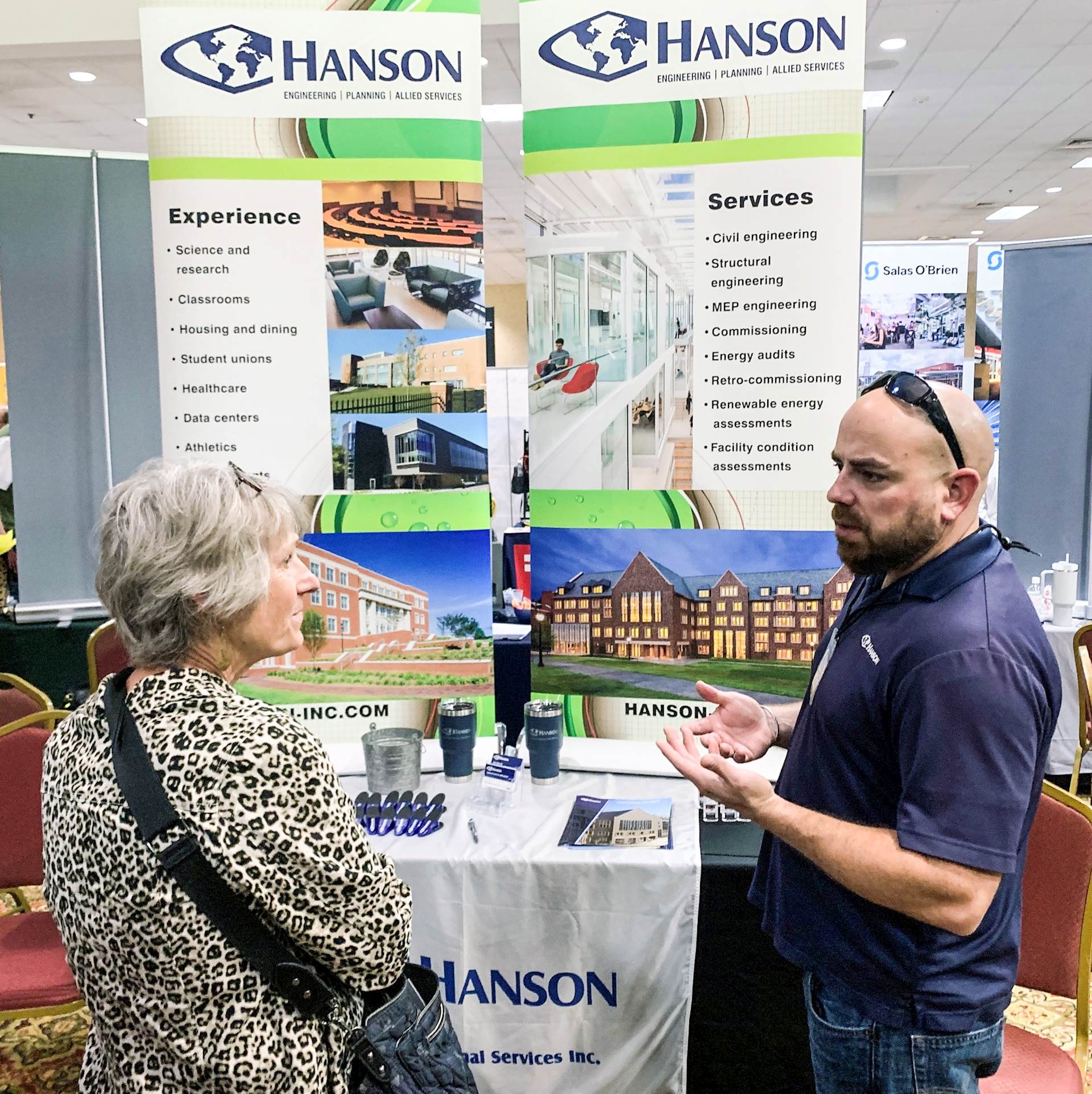Two people stand in front of Hanson’s table and banners in a hotel event space; person on right is gesturing while talking to the other person