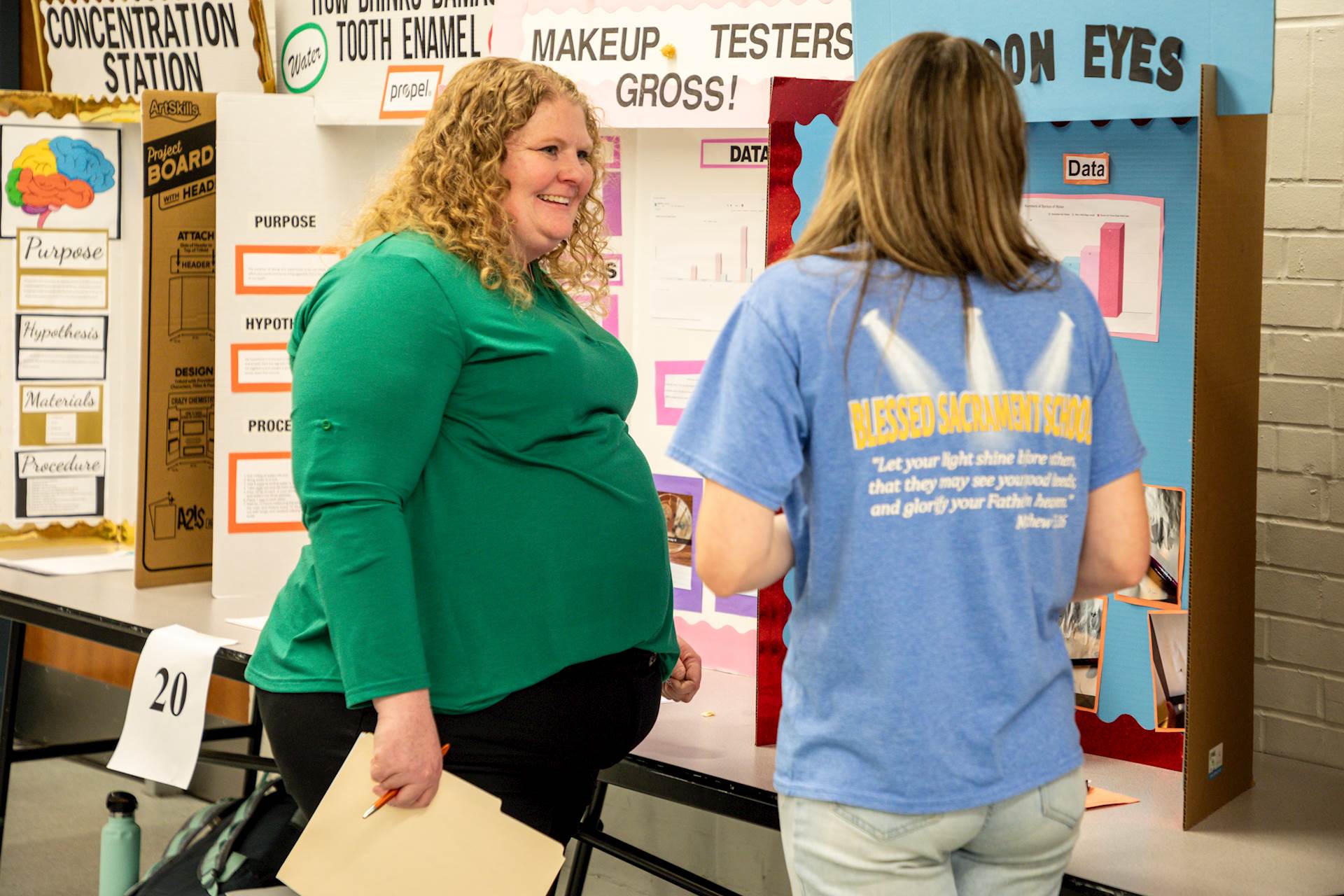 Woman in green shirt and black pants talks to student in jeans and blue T-shirt in front of science fair display