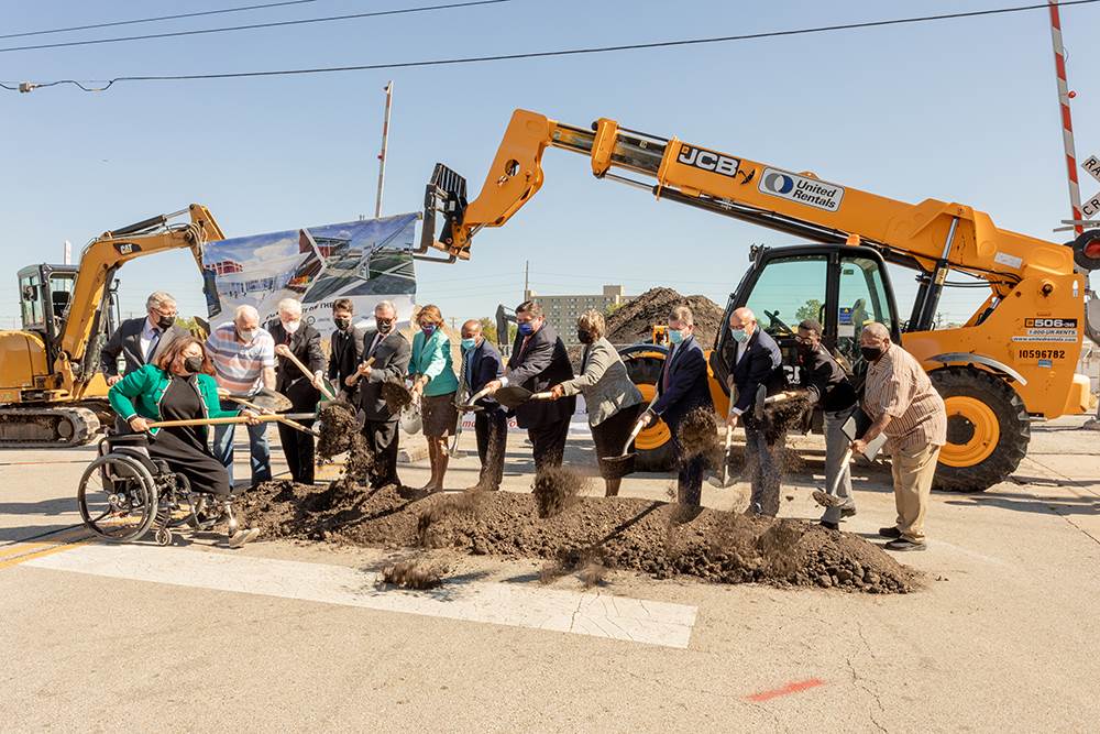 Fourteen people toss dirt with shovels in front of construction equipment displaying a banner at a groundbreaking ceremony on