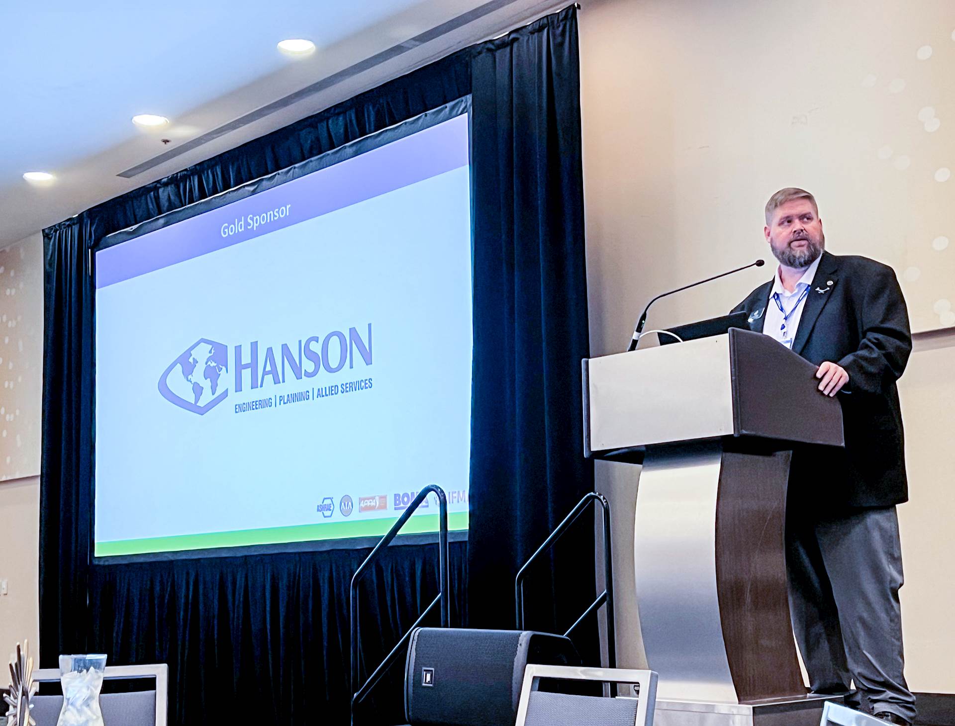Person stands behind a lectern next to a project screen in a meeting room; screen shows Hanson logo on presentation slide for conference sponsors.