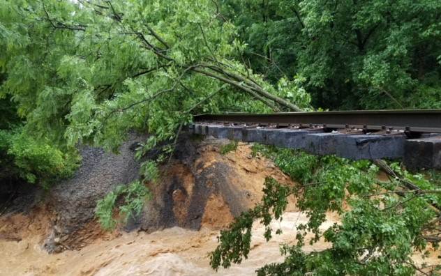 A fallen tree lies on a rail bridge as water rushes below it. 