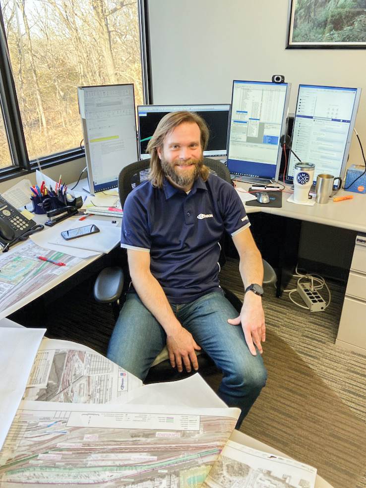 Andy Rucker sits in a chair at his desk, smiling at camera, with railway facility maps in front of him