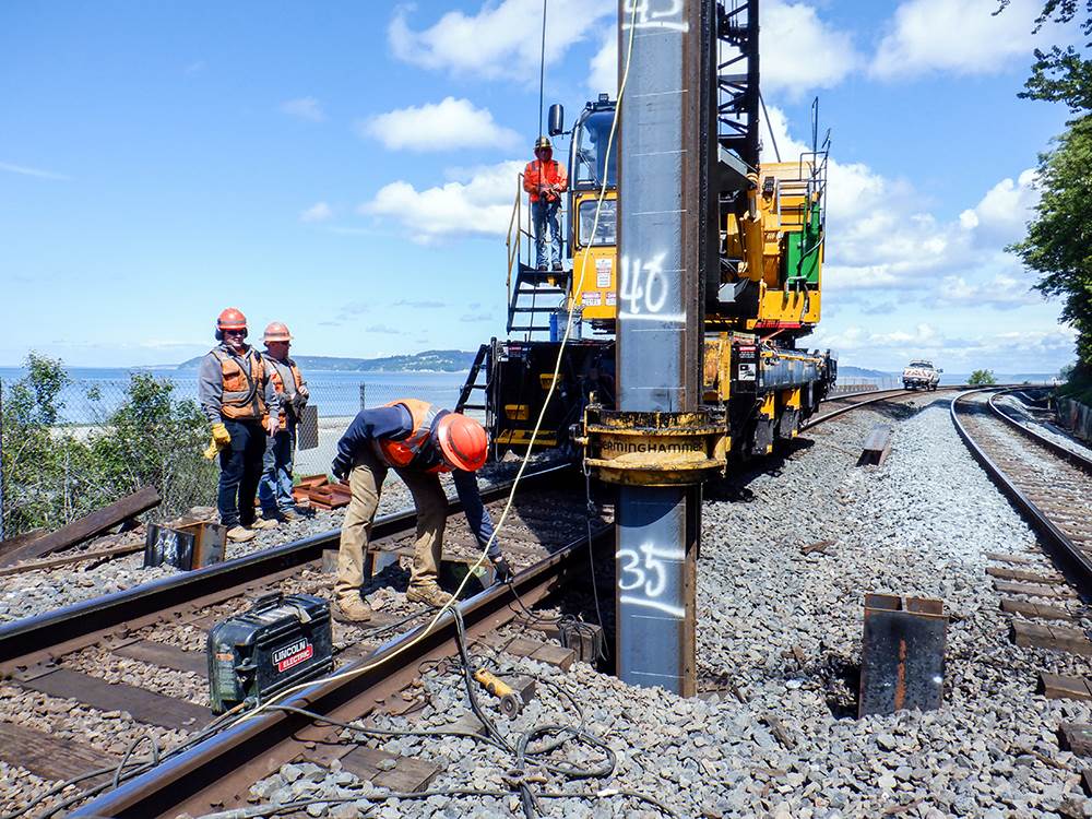 Four workers use a pile driving hammer on a double-track rail bridge; a beach and waterway is behind them