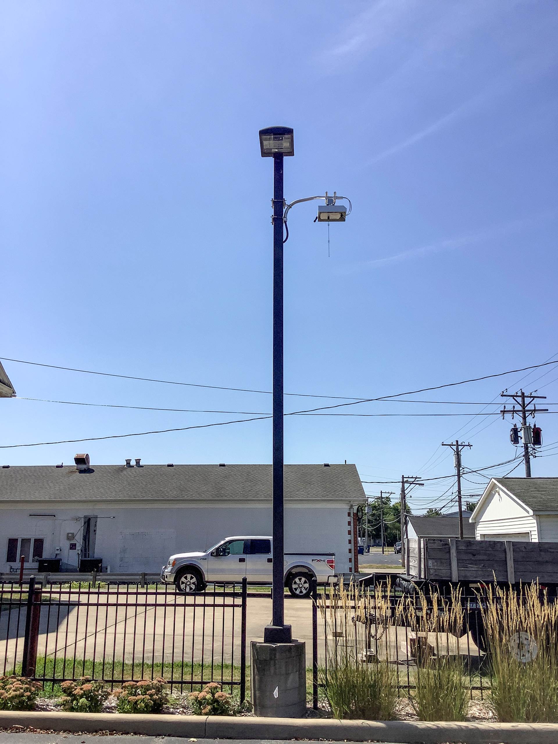 A light pole with a data collection device at the top. The light pole is between two parking lots, with a small commercial building and an alley in the background.