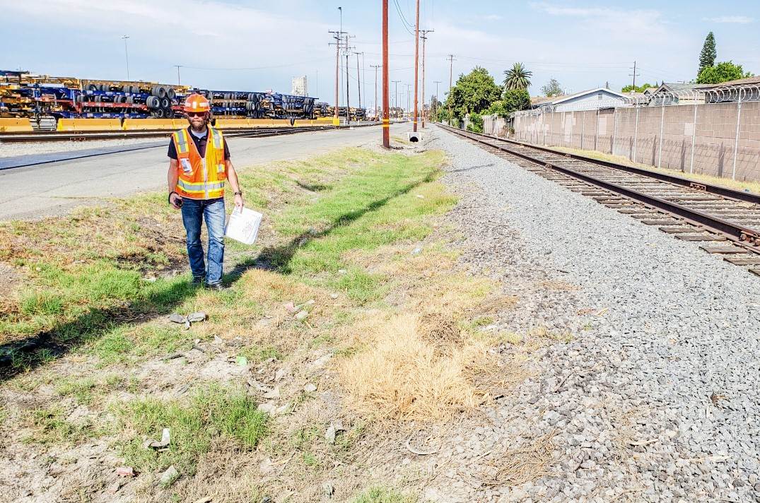 Andy Rucker, holding project documents, stands in a grassy area of a railroad’s intermodal terminal on a sunny day