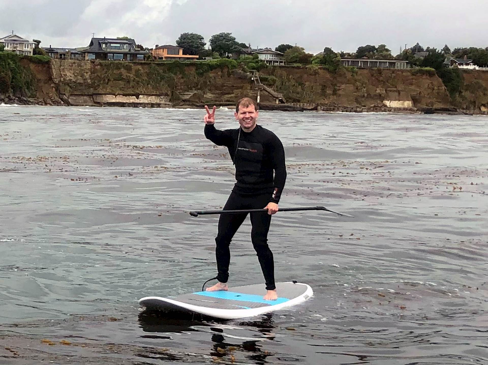 Matt Willey, wearing a wetsuit, gives a “peace” hand sign and stands on a paddleboard in the waters of Monterey Bay while hol