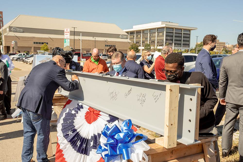 People use permanent markers to sign a steel beam on a sunny day, surrounded by a small crowd