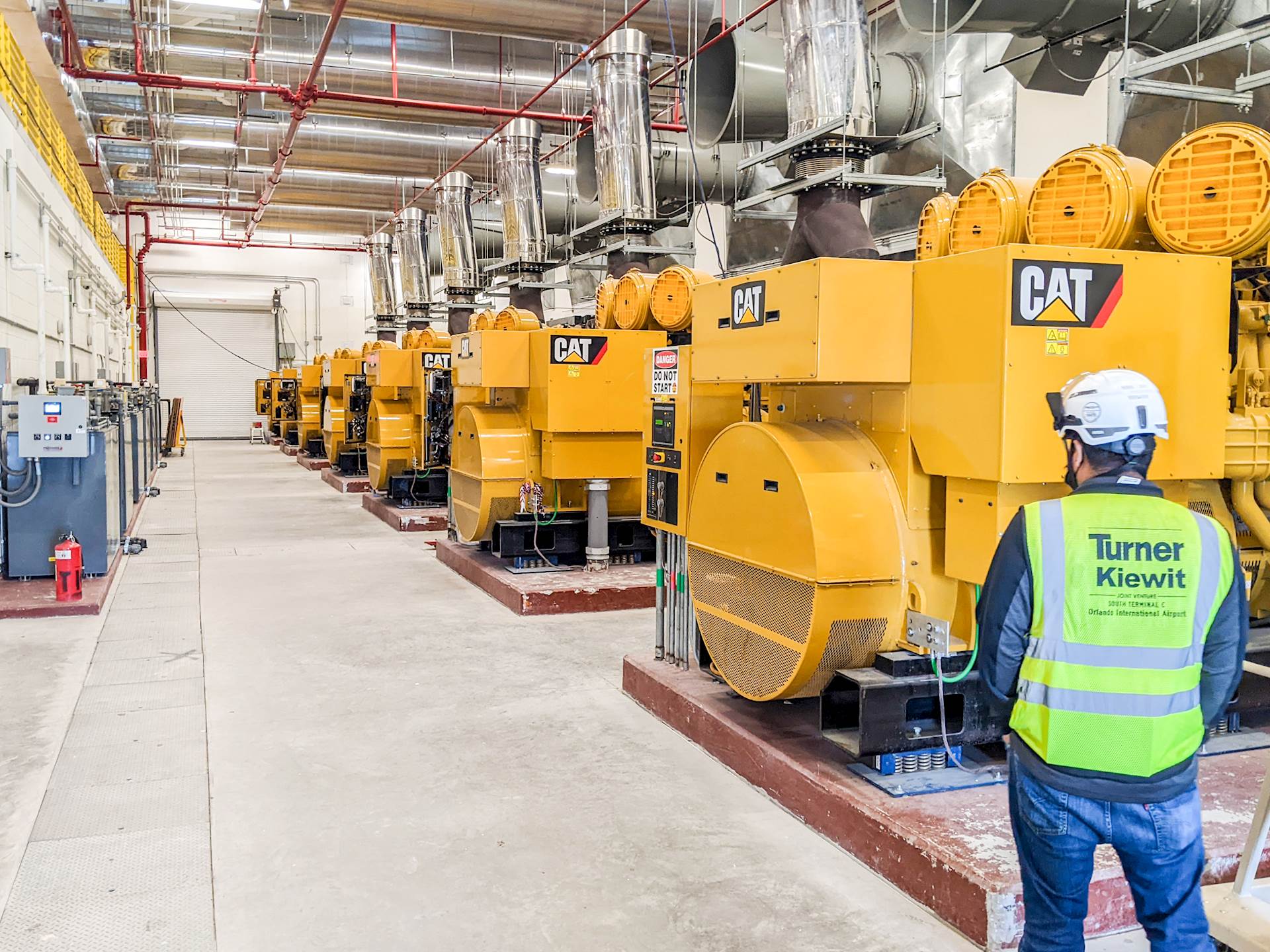 A person in a hard hat and safety vest stands in front of a row of several large generators inside a large mechanical area