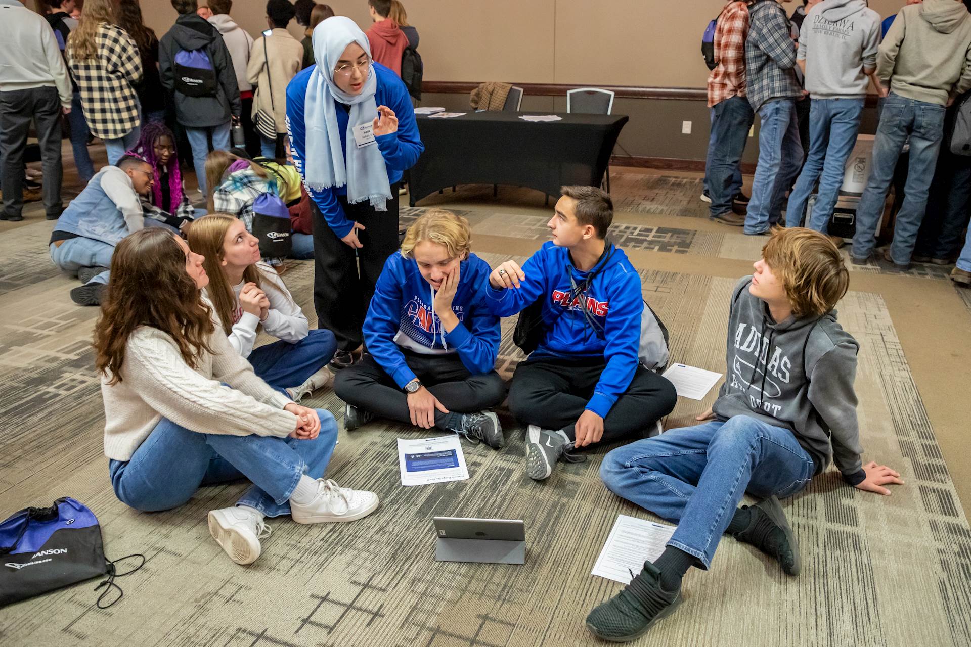 Hanson employee speaks to five students sitting on the floor of a conference room with beige, patterned carpet