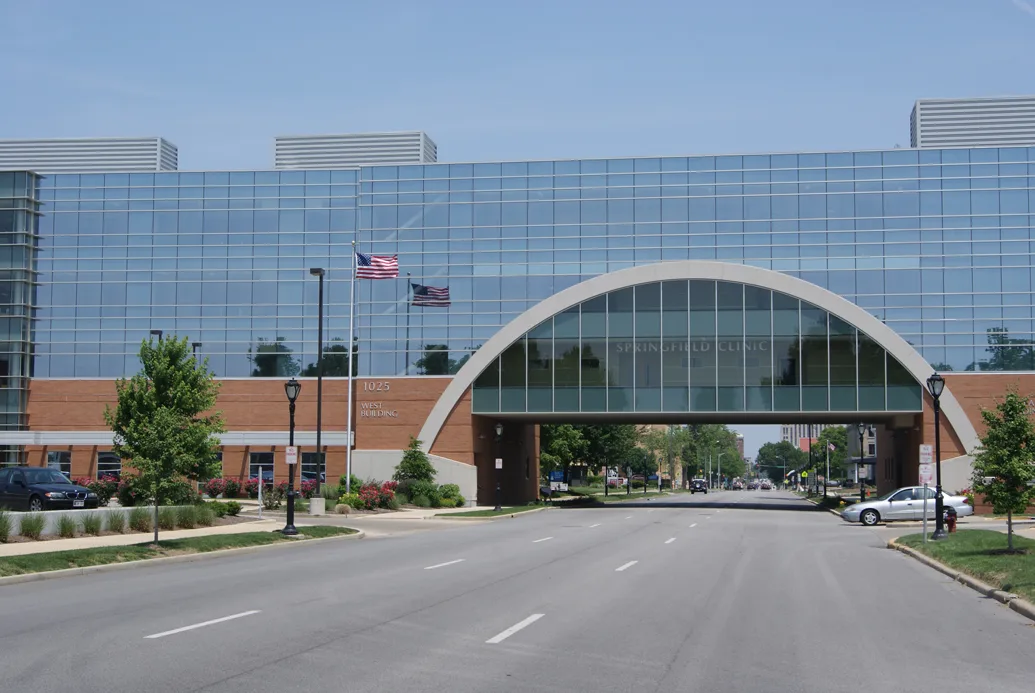 Photo of medical office building with three-story enclosed pedestrian bridge and roadway passing underneath. Silver car pulling out of parking lot at right and U.S. flag flying at left.