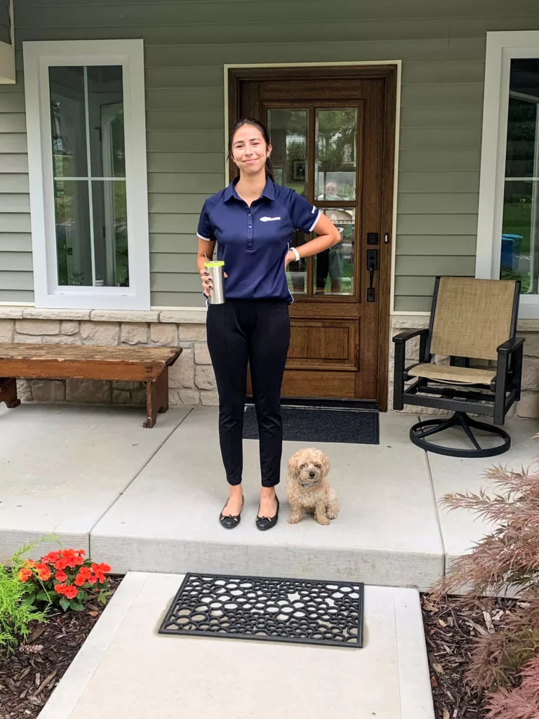 Woman with dark hair pulled back, standing on front porch of house with green siding, wearing black pants and a navy blue short-sleeved polo shirt with a small dog by her side. 