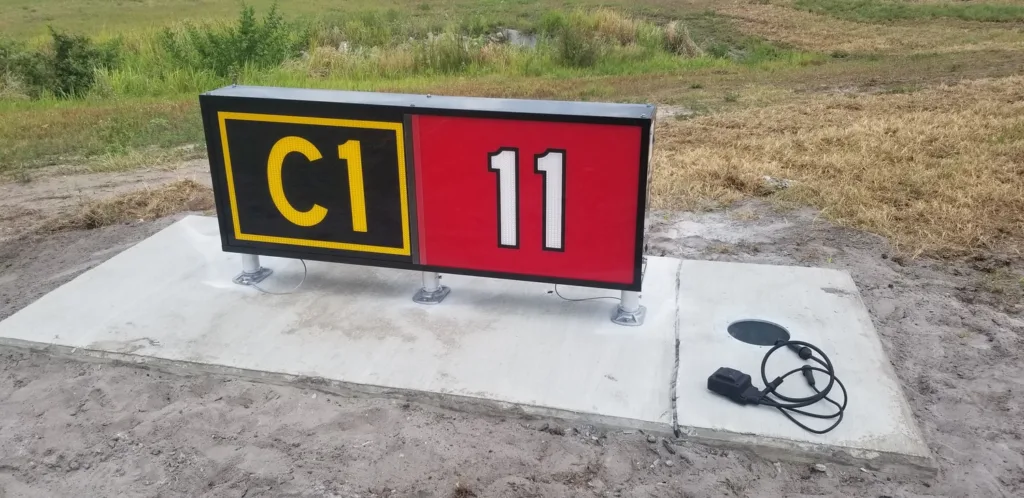 Red, black and yellow airfield sign on concrete slab with grass in background