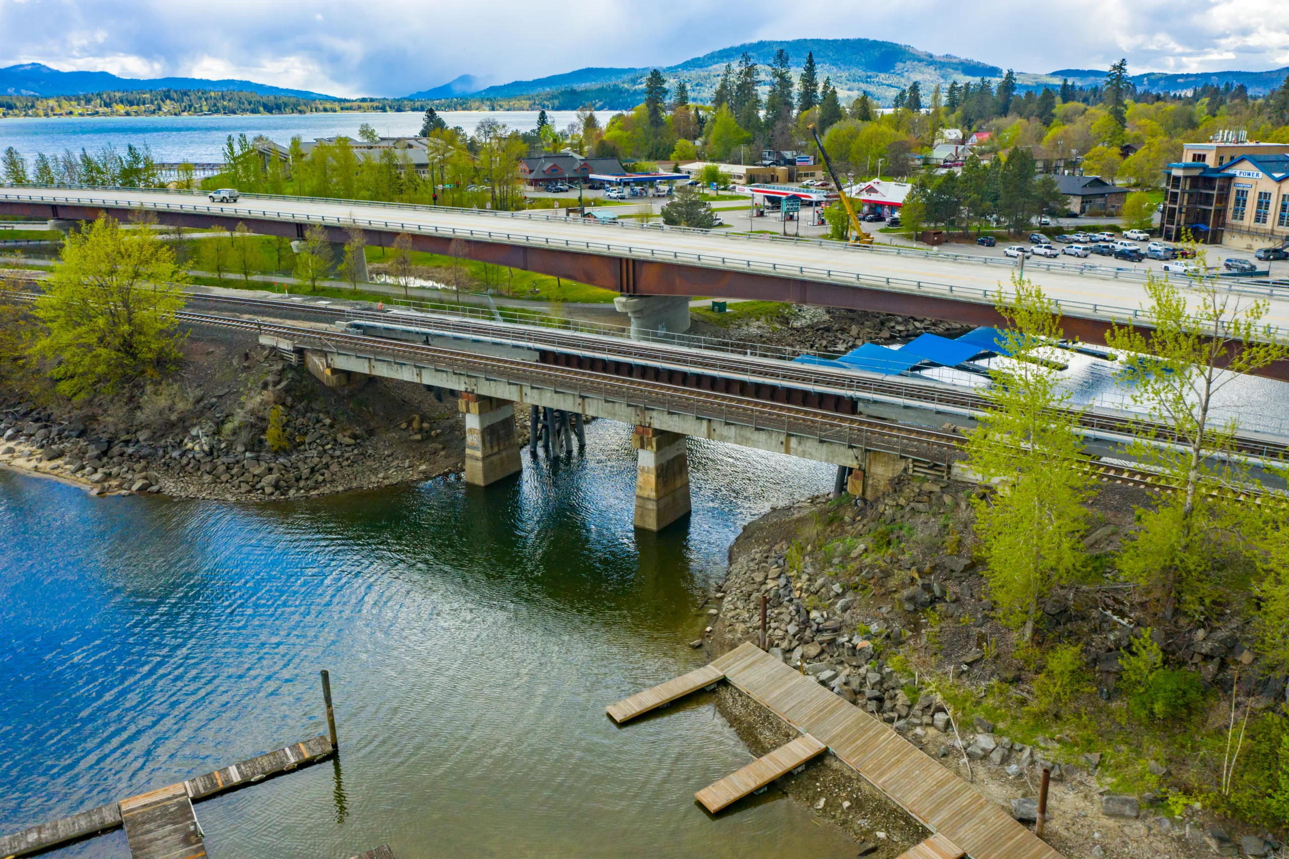 Aerial view of a railroad track side by side with a roadway going over a river with trees and mountains in the background
