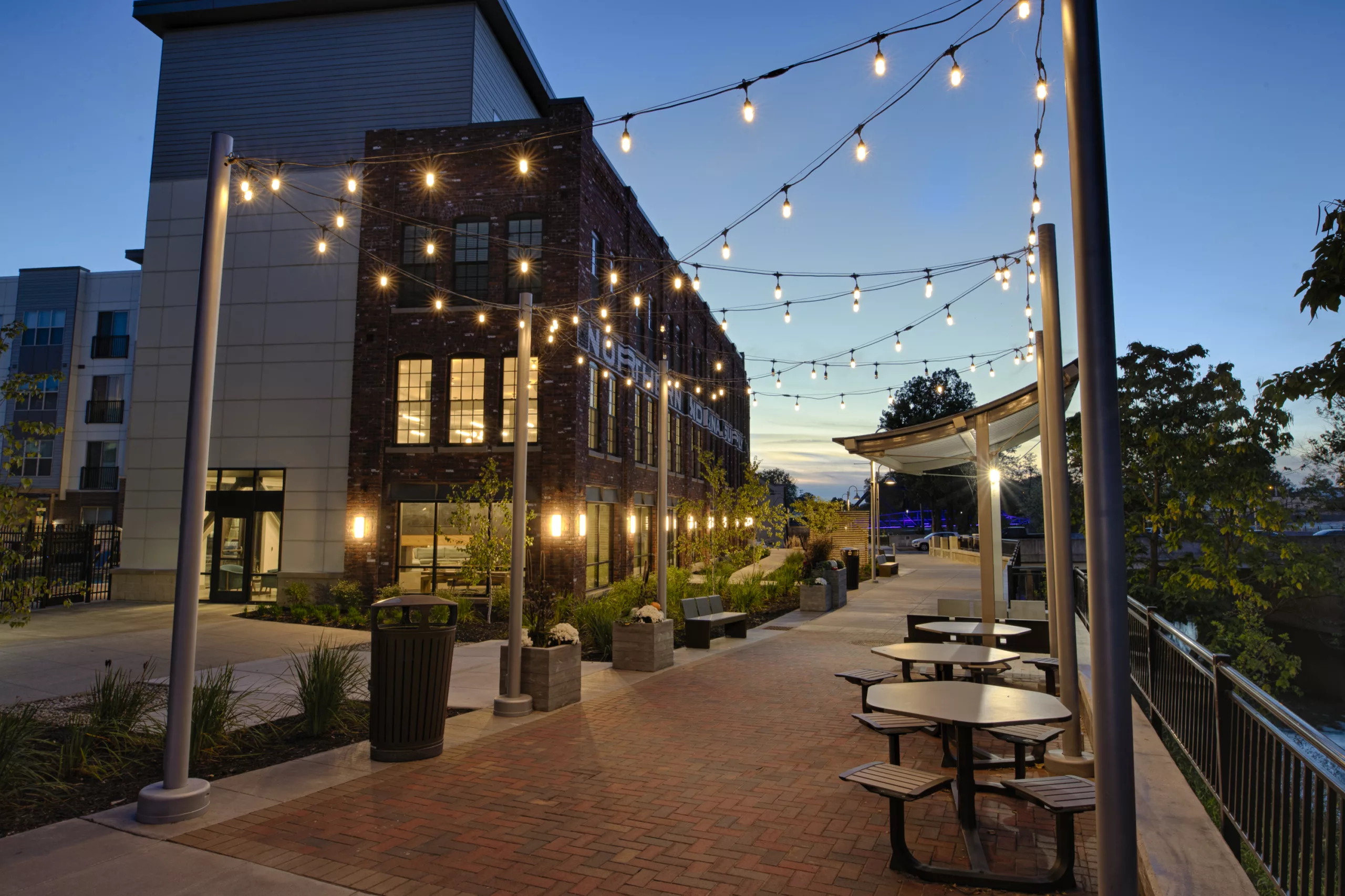 Dusk view of buildings with decorative lighting, tables and brick path