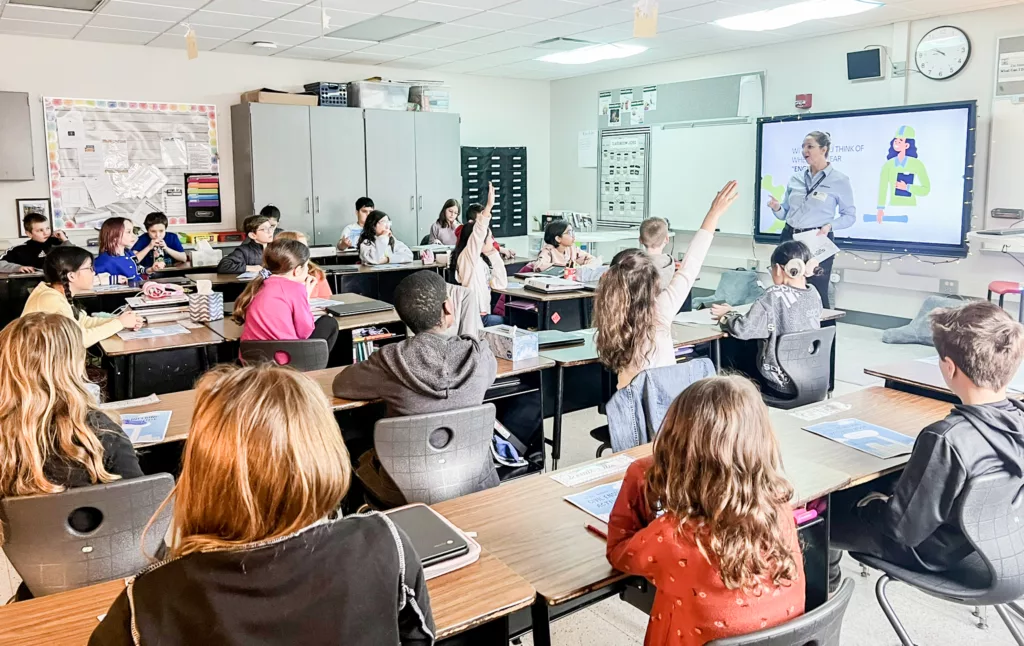 Children in classroom raising their hands while woman at the right speaks