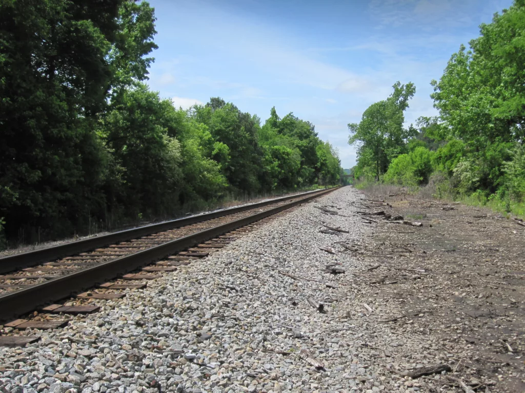 Railroad tracks with gravel in foreground, blue sky and green trees in foreground and background