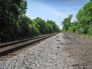 Railroad tracks with gravel in foreground, blue sky and green trees in foreground and background