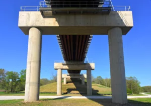 View of underside of bridge length with concrete piers and blue sky and green grass in background