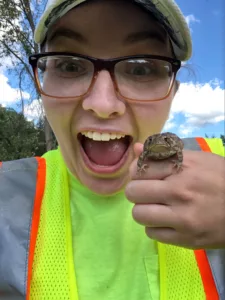 Woman wearing glasses, a cap and a neon-yellow safety vest, smiling, holding a small frog