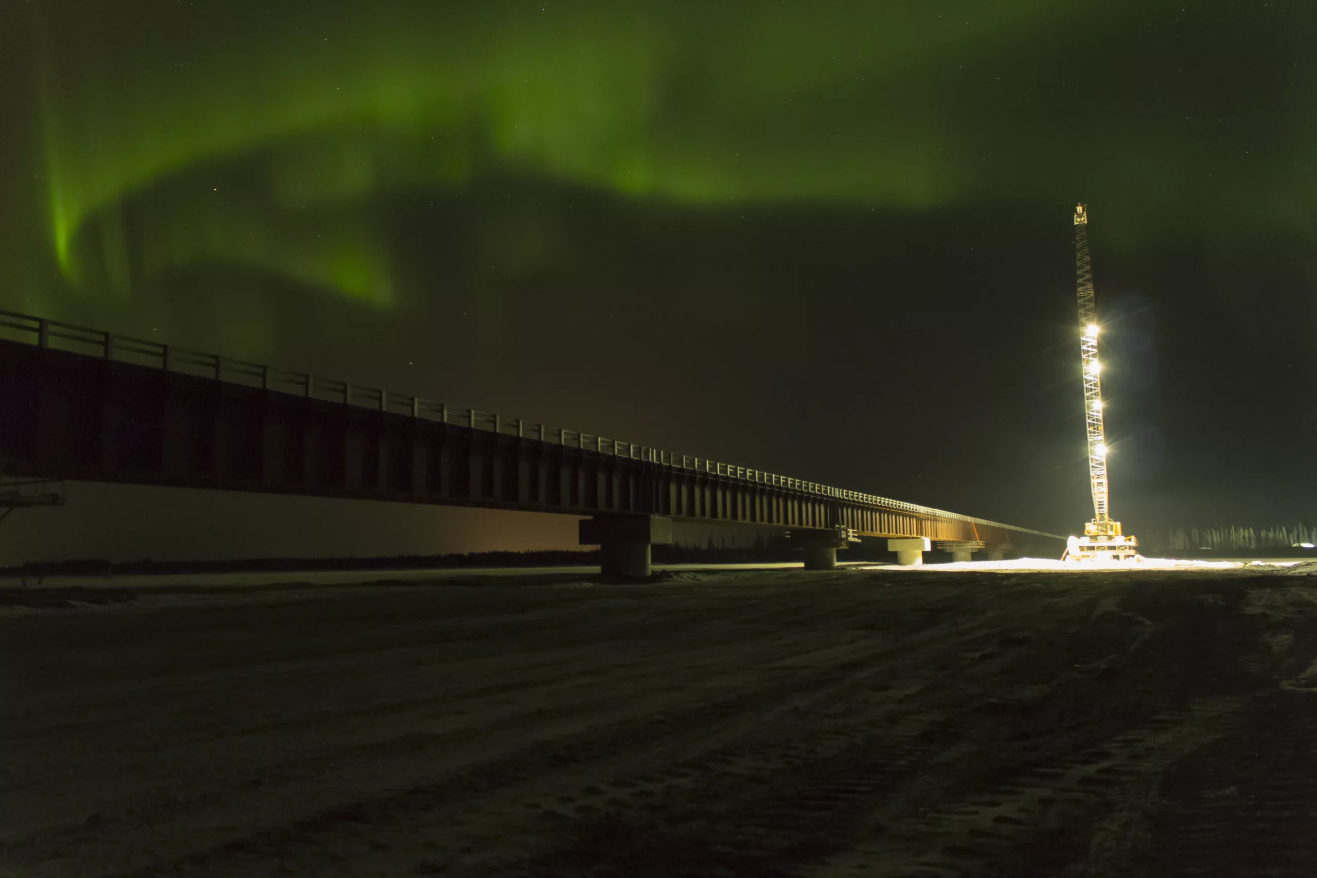 Long bridge over frozen river at night with northern lights above and lit-up tower to the right.