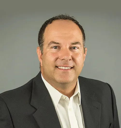 Man with short, dark hair wearing black jacket and white button-down, collared shirt on light beige background