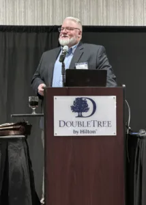 Man standing behind lectern, wearing dark suit jacket with light blue button-down shirt, with black backdrop.