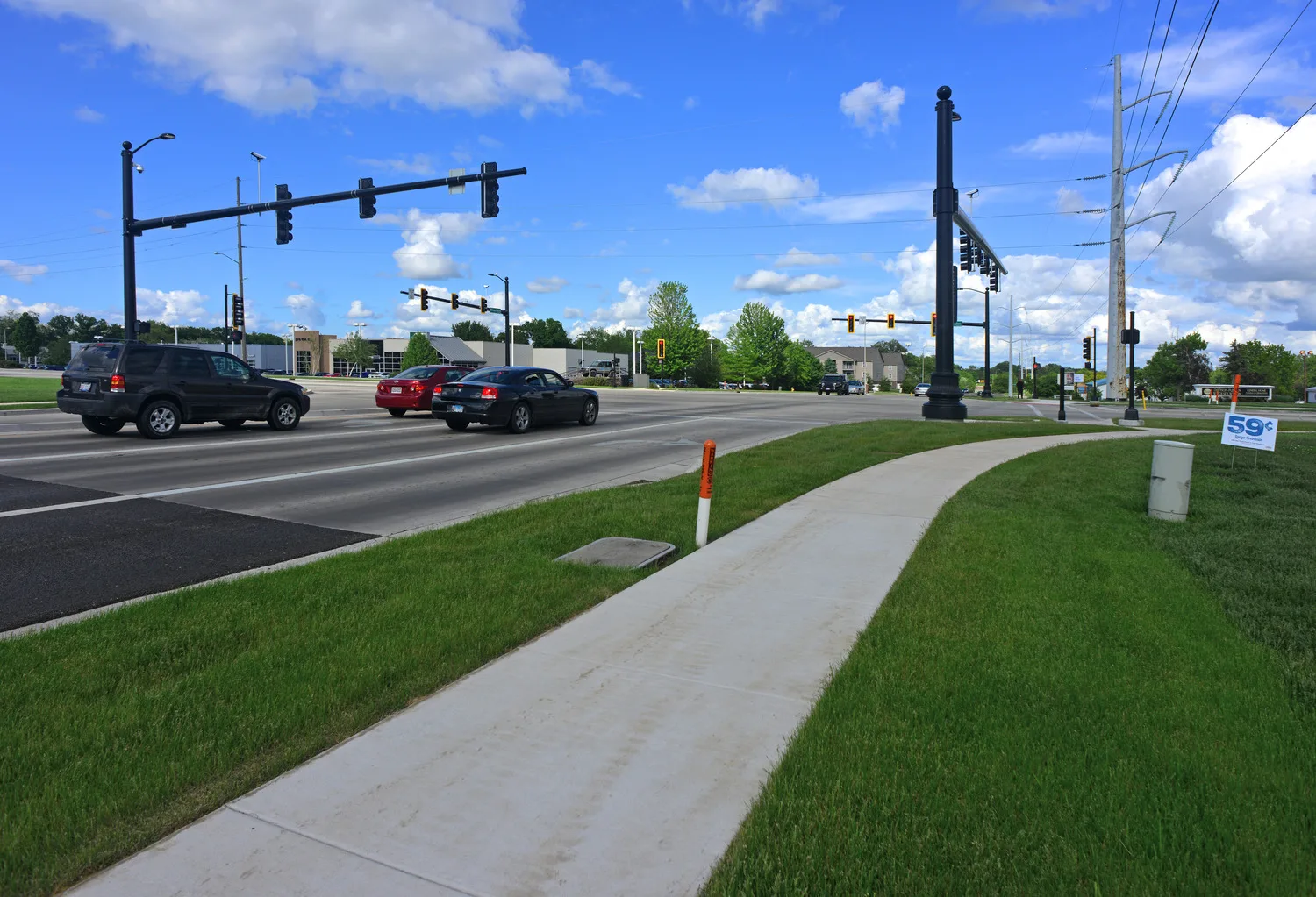 Streetside view of newly updated roadway with cars at intersection featuring black traffic lights and sidewalk in foreground