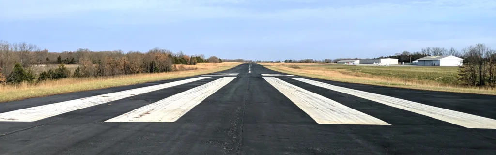 Black paved runway with white stripes and blue sky in background; hangars off to right and foliage off to left