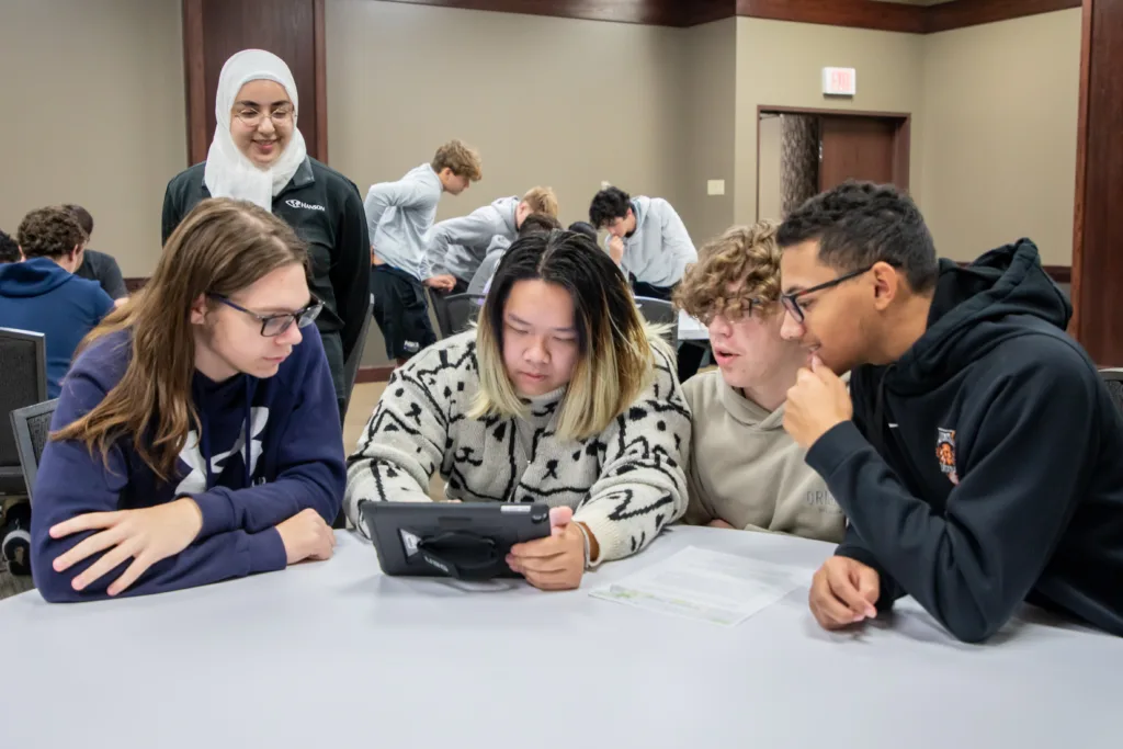 Four students sitting at table looking at tablet, with Hanson employee smiling in background and other students at tables also in background