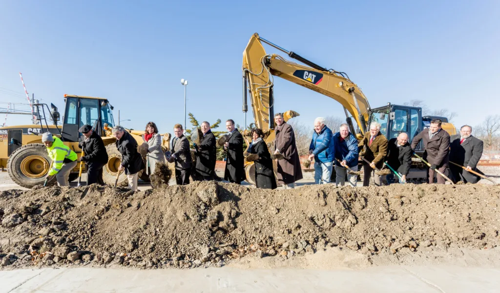 Line of people with shovels standing on dirt near construction implement at site of Ash Street underpass groundbreaking