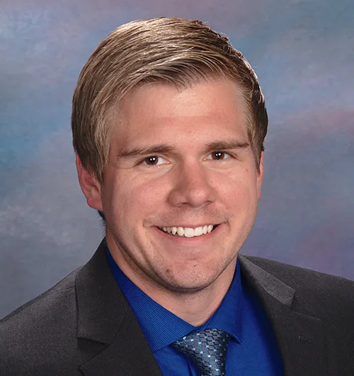 Man with dark blond hair, wearing dark suit jacket with royal blue shirt and patterned blue tie against blue and purple background