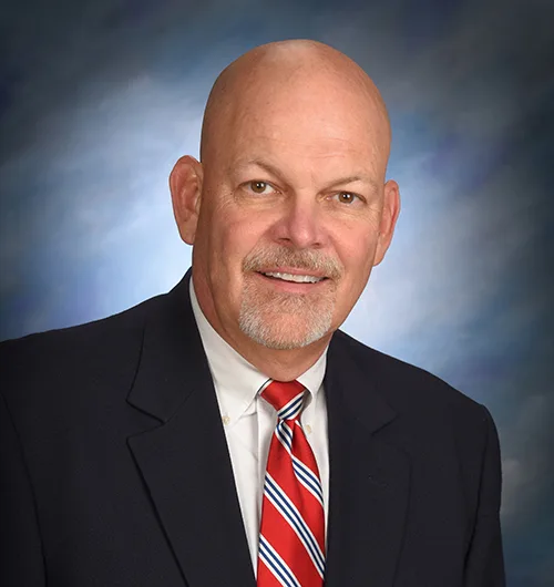 Bald man with beard and mustache wearing dark suit jacket, white button down, collared shirt and red, blue and white striped tie