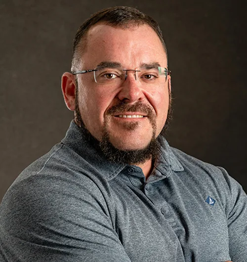 Man with dark hair, beard and moustache, wearing gray shirt with dark background