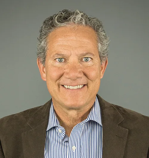 Man with gray, curly hair wearing down brown sport coat with blue and white striped button down, collared shirt on light beige background