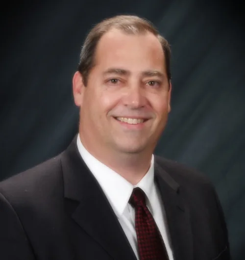 Man with dark hair against dark background, wearing black suit jacket with white button down, collared shirt and red and black checked tie