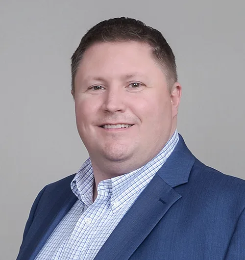 Man with short dark hair wearing blue suit jacket with white and blue checked button down, collared shirt against beige background