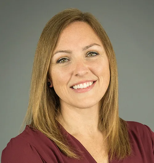 Woman with straight brown hair to shoulders, smiling, wearing maroon top with dark beige background