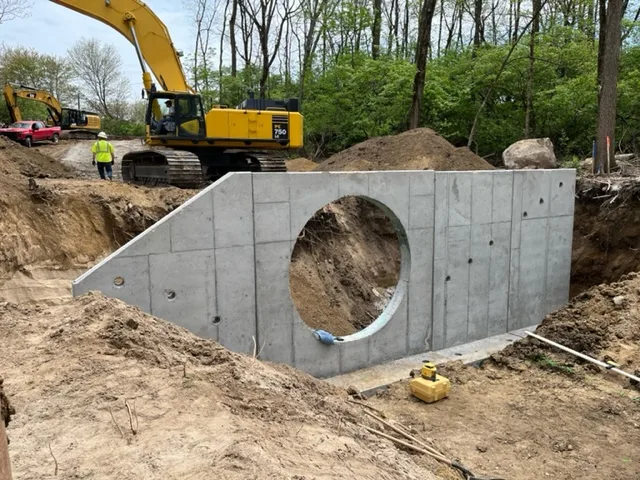 Concrete wall in dirt at construction site with equipment, worker, red truck and trees in background