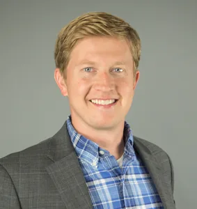 Man with blond hair wearing gray suit jacket with light checked pattern and blue plaid shirt with light beige background