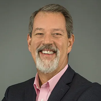 Man with gray hair and gray facial hair with open-mouthed smile, wearing pink button-down, collared shirt and black jacket with dark beige background