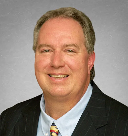 Man with short blond hair wearing black pin-striped jacket with light blue button-down shirt and yellow, red and blue tie and light gray background
