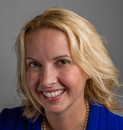 Woman with blond hair wearing pearl necklace and royal blue jacket in front of gray background