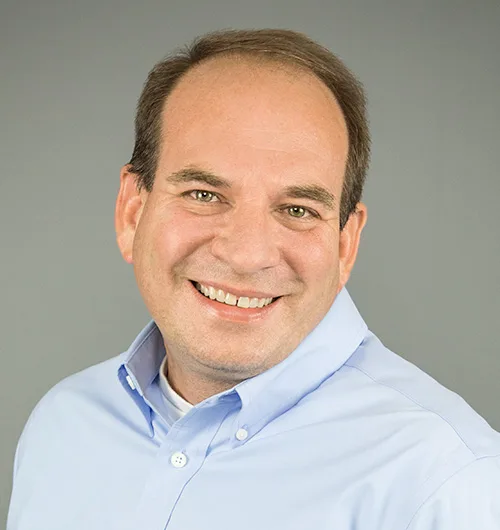 Man with light brown hair wearing light blue button down, collared shirt with head tilted to the right on light beige background