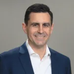 Man with black hair, wearing dark blue jacket over white button down, collared shirt, against beige background