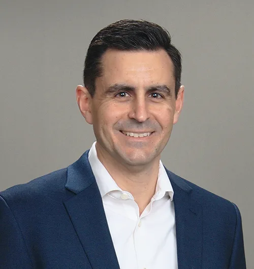 Man with black hair, wearing dark blue jacket over white button down, collared shirt, against beige background
