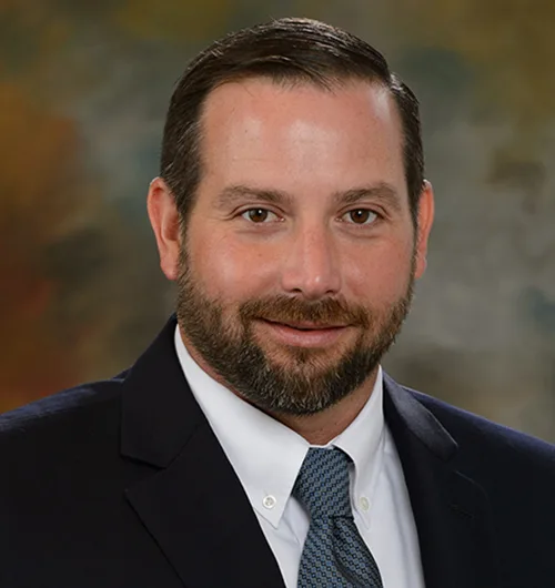 Man with short brown hair and brown facial hair, brown eyes, wearing navy blue suit jacket, white collared button-down shirt and blue patterned tie with marbled brown background