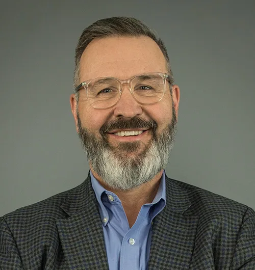 Smiling man with dark hair and graying beard and mustache wearing glasses and checked sport coat with light blue button-down, collared shirt against dark beige background