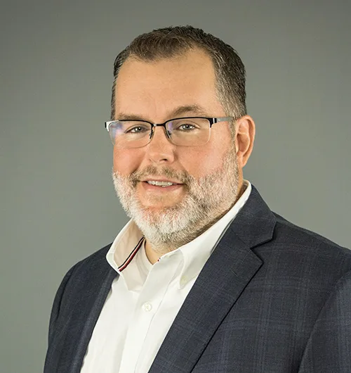Man with dark hair, glasses and white beard and mustache wearing dark suit jacket with light check pattern and white collared, button-down shirt with light beige background